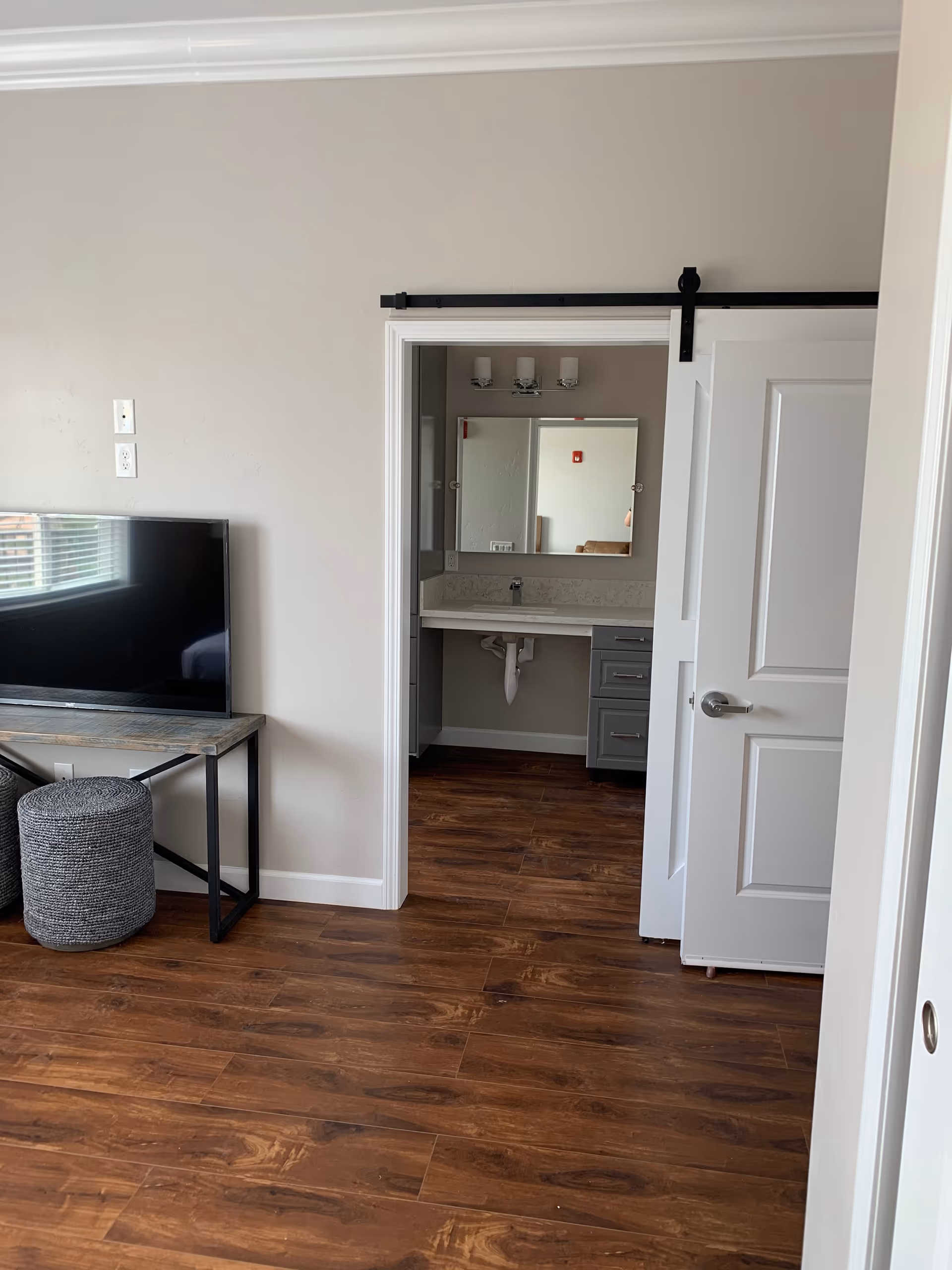 Interior view of a room with wooden flooring, a flat-screen TV on a wooden and metal stand, and a gray pouf. A sliding barn door is partially open, revealing a bathroom sink with a mirror and light fixtures above it.