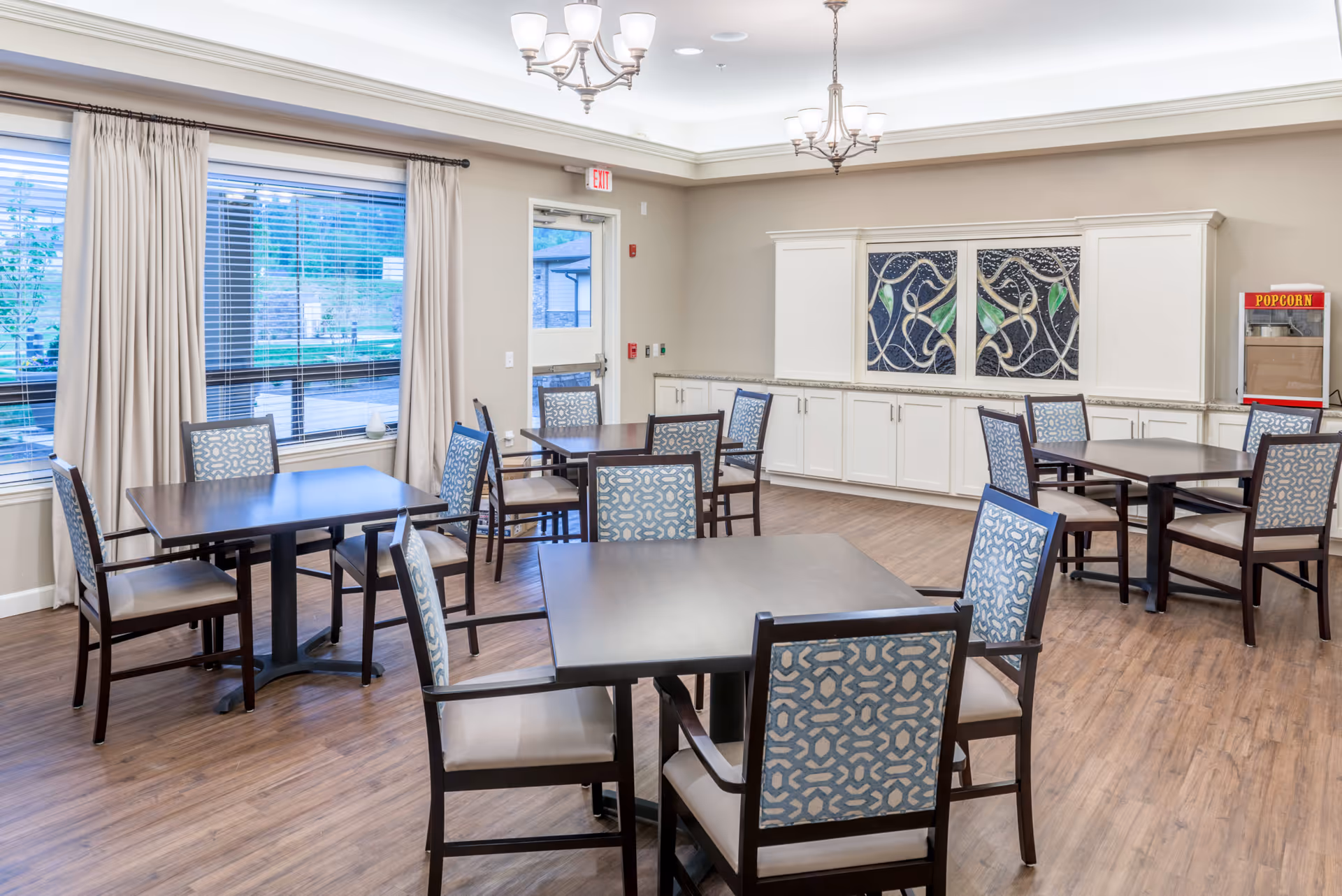 A bright dining room with several dark wood tables and patterned cushioned chairs arranged neatly on a wood floor. Large windows with beige curtains allow natural light to enter. A popcorn machine is visible in the corner near white cabinetry with decorative panels above.