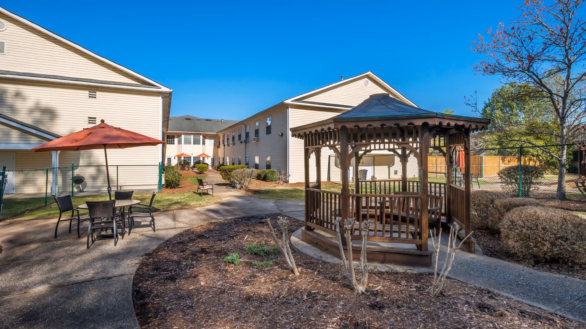 Outdoor courtyard area of a senior living facility with a wooden gazebo, patio table with chairs and an umbrella, surrounded by bushes and trees, with beige multi-story buildings in the background under a clear blue sky.