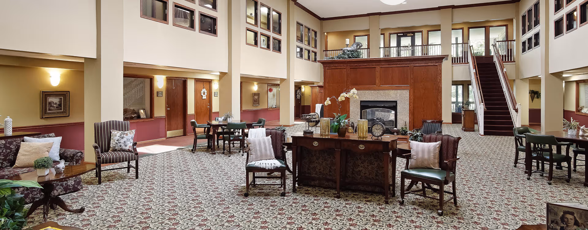 Spacious senior living facility common area with patterned carpet, multiple seating arrangements including armchairs and tables, a large wooden fireplace centerpiece, and a staircase leading to an upper floor. The walls have framed pictures and windows overlooking the area.