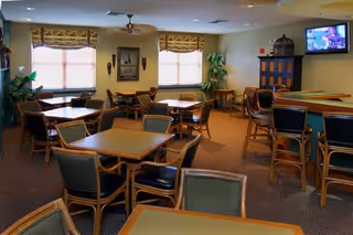 A common area with multiple square tables and chairs arranged for seating. The room has beige walls, carpeted floor, two windows with patterned valances, a ceiling fan, potted plants, a large cabinet, and a wall-mounted TV showing a program.