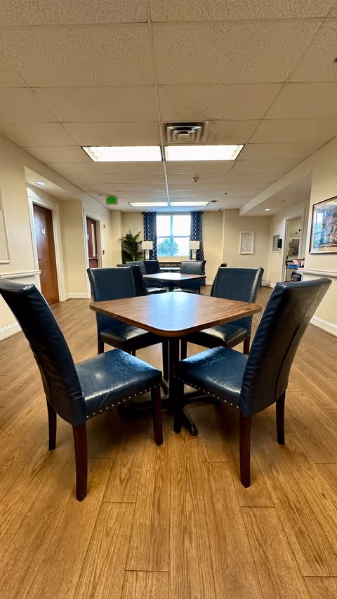 Interior view of a common area in a senior living facility with a wooden table surrounded by four dark blue cushioned chairs. The room has wood flooring, beige walls, and a ceiling with fluorescent lights. In the background, there are more tables and chairs near a window with blue patterned curtains, a potted plant, and two lamps on a side table.