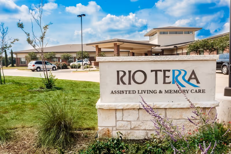 Exterior view of Rio Terra Senior Living facility with a stone sign in the foreground that reads 'RIO TERRA ASSISTED LIVING & MEMORY CARE'. The building and parking area with cars are visible in the background under a partly cloudy blue sky.