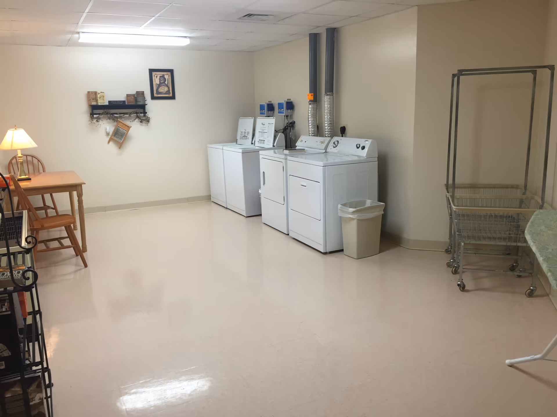 A laundry room with two washing machines and two dryers along the right wall, a trash bin next to the machines, a metal laundry cart, a wooden table with a lamp and chairs on the left side, and a small shelf with decorative items on the wall.
