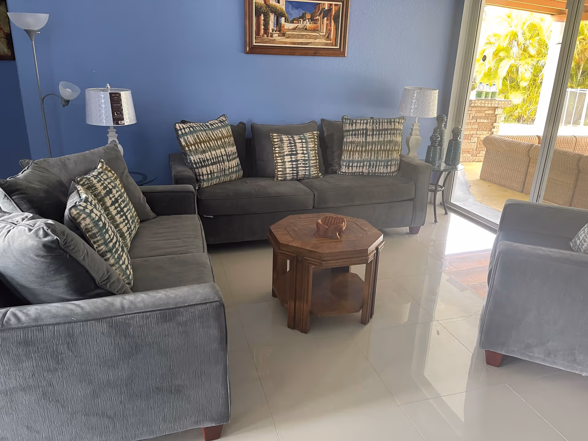 A cozy living room with two gray sofas and one gray armchair arranged around a wooden octagonal coffee table. The sofas have patterned cushions. There are two white table lamps on either side of the central sofa, and a floor lamp is visible on the left. A framed painting hangs on a blue wall behind the sofa. Large sliding glass doors open to an outdoor patio area with wicker furniture and lush greenery.