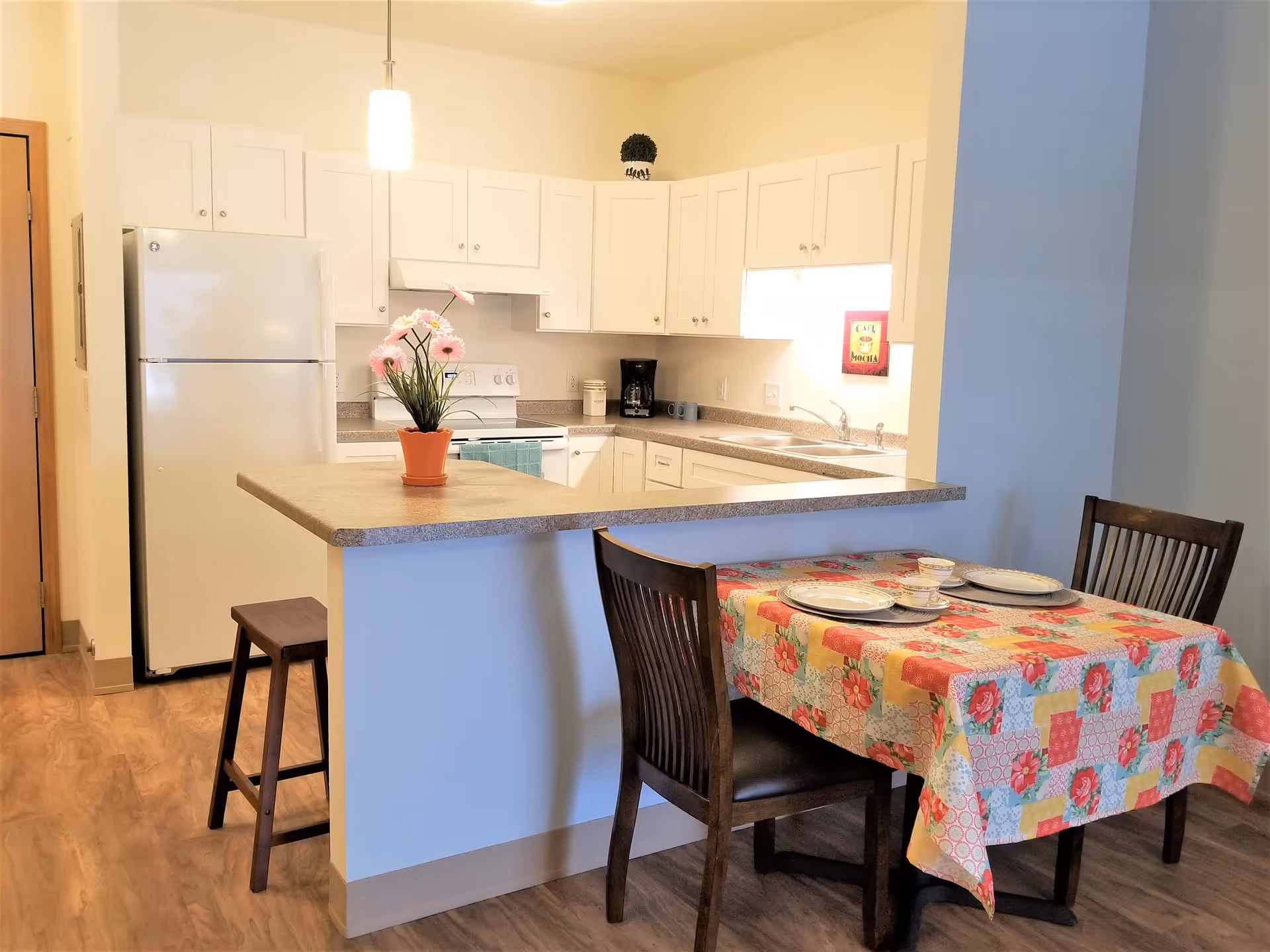 Open white kitchen with a breakfast bar topped by a potted flowering plant and a nearby dining table set with a colorful tablecloth.