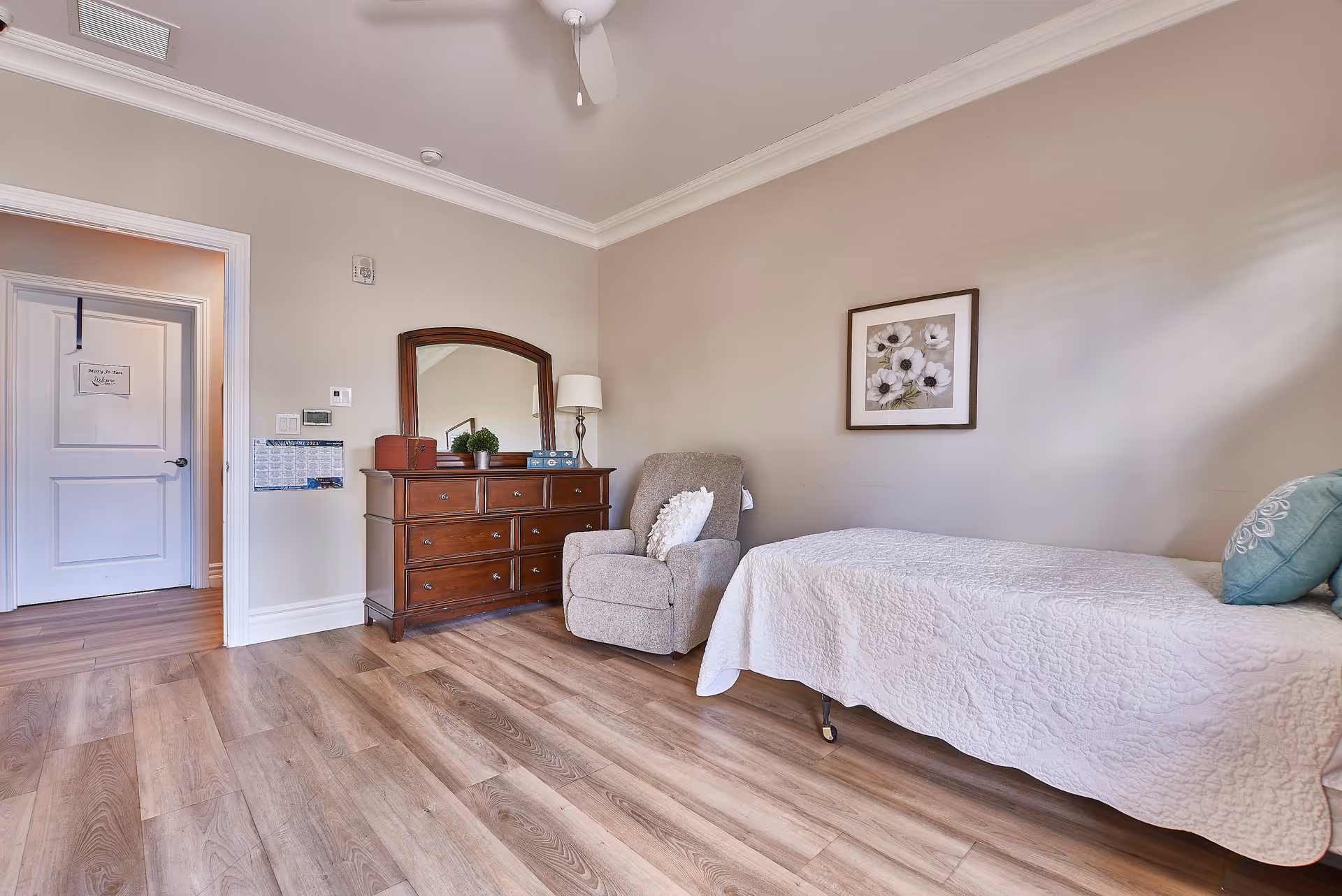 A cozy bedroom with a single bed covered in a white quilt, a gray upholstered armchair with a white pillow, and a wooden dresser with a mirror, lamp, and decorative items. The room has light beige walls, wood flooring, a ceiling fan, and a framed floral picture on the wall.