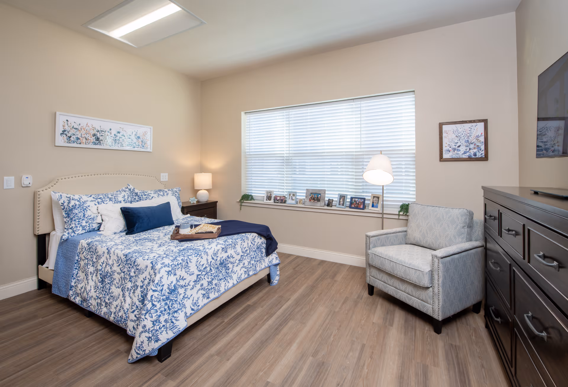 A cozy bedroom with a bed featuring blue and white floral bedding, a beige upholstered headboard, and a tray with a cup and book on the bed. There is a nightstand with a lamp beside the bed, a large window with blinds and several framed photos on the windowsill. A patterned armchair and a dark wooden dresser with a TV on top are also visible. The walls are beige and the floor is wood.