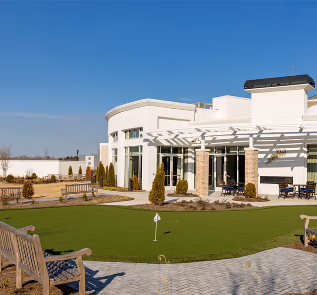 Modern white senior living building with a small putting green, benches, and patio seating under a pergola in a landscaped courtyard.