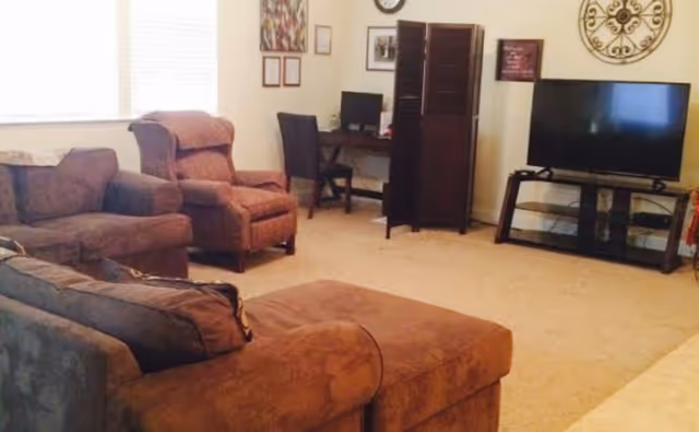 A cozy living room with brown upholstered furniture including a sofa, armchair, and ottoman. There is a flat-screen TV on a black stand against the wall, a small desk with a computer and chair, and decorative wall art including a clock and framed pictures. The room is carpeted and well-lit by natural light from a window with blinds.
