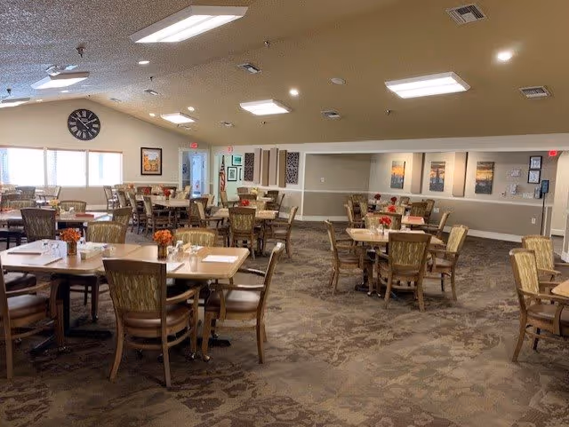 A spacious dining room with multiple wooden tables and chairs arranged neatly. Each table has a small floral centerpiece and place settings. The room has a beige carpet with a patterned design, light-colored walls, and several framed pictures on the walls. Ceiling lights provide bright illumination, and a large clock is visible on one wall near windows.