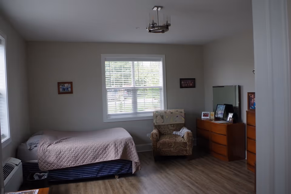 Simple bedroom with a single bed, upholstered armchair, wooden dressers and a window letting in daylight.
