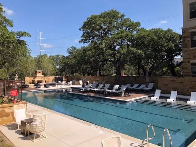 Outdoor swimming pool area with several white lounge chairs lined up along the poolside. There are trees and a stone wall in the background, along with a fireplace and additional seating. The sky is clear and blue.