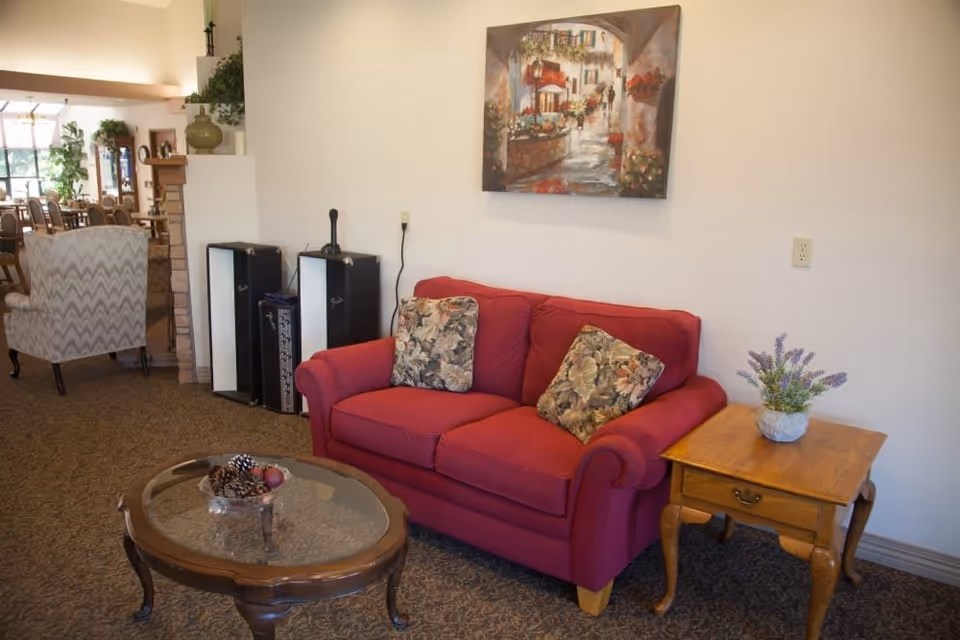 A cozy living room area featuring a red loveseat with two floral cushions, a wooden side table with a potted lavender plant, and a glass-top coffee table with decorative pine cones and fruit. A painting of a street scene hangs on the wall above the loveseat. In the background, there is a patterned armchair and a dining area with tables and chairs, along with some plants and decorative items.