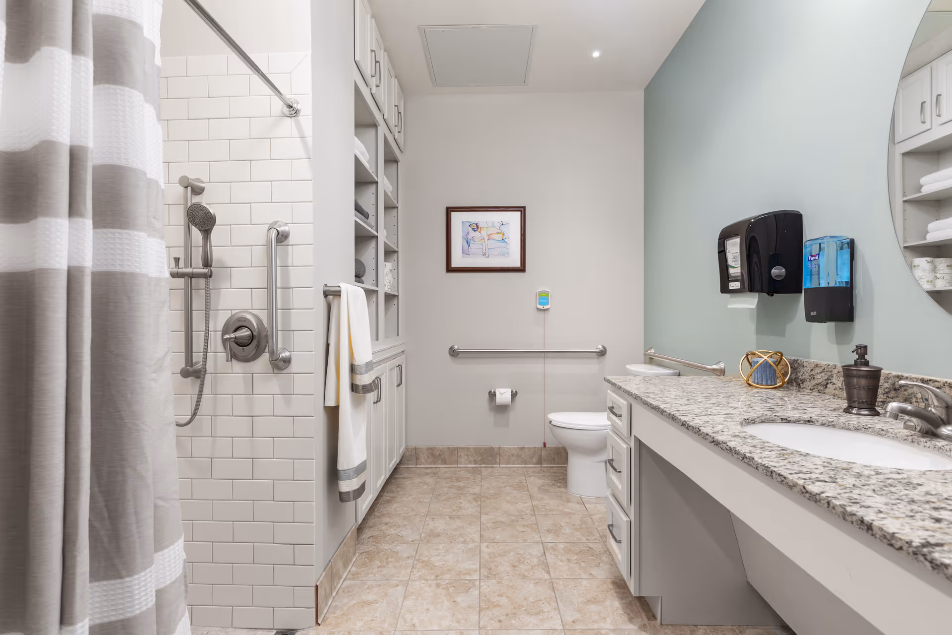 A clean and spacious bathroom with beige tiled floor, a shower area with white subway tiles and a striped shower curtain on the left, a white toilet with a grab bar on the back wall, and a long granite countertop with a sink, soap dispenser, and paper towel dispenser on the right. There are built-in shelves with towels and other items, a framed picture on the wall above the toilet, and a light blue accent wall behind the sink.