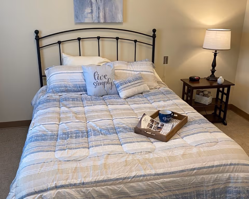 Neatly made bed with blue-and-white striped bedding, a decorative 'live simply' pillow, a tray with a mug on the bed, and a nightstand with a lamp.
