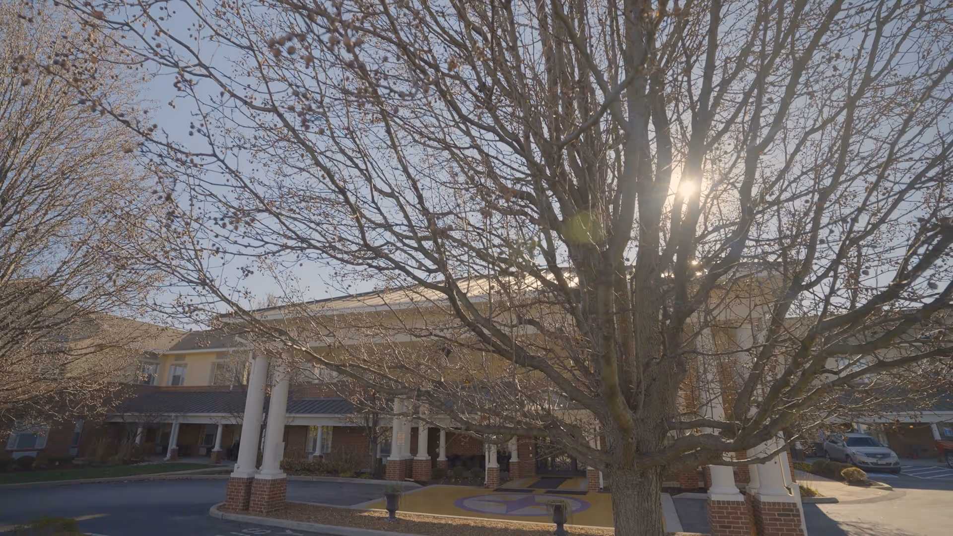 Exterior view of a senior living facility building with a covered entrance supported by white columns. Leafless trees are in the foreground, and the sun is shining through the branches. Cars are parked near the building under a clear sky.