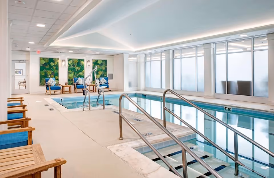 Bright indoor lap pool with metal handrails, seating area, and large frosted windows.
