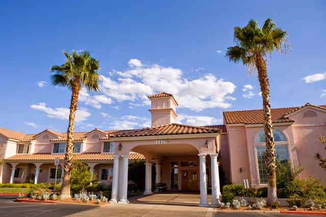Exterior view of a senior living facility with a covered entrance supported by white columns, two tall palm trees, and a clear blue sky with scattered clouds. The building has a tiled roof and large windows.
