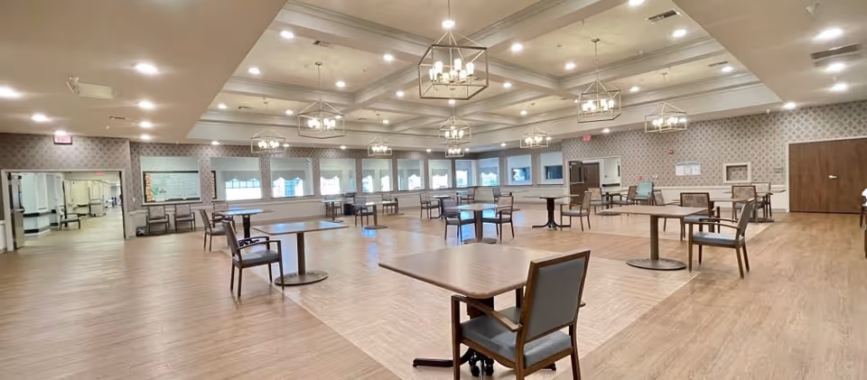 A spacious dining room with multiple wooden tables and chairs arranged neatly on a wood-patterned floor. The room features patterned wallpaper, several large windows, and multiple ceiling light fixtures with chandeliers. There are doorways visible on the far walls leading to other areas of the facility.