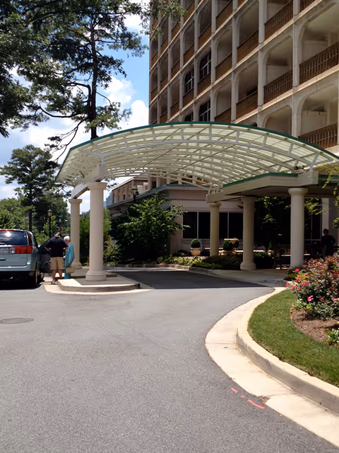 Entrance area of a multi-story building with a covered driveway supported by columns. Two people are standing near a parked vehicle under the canopy. Trees and bushes surround the driveway, and the sky is partly cloudy.