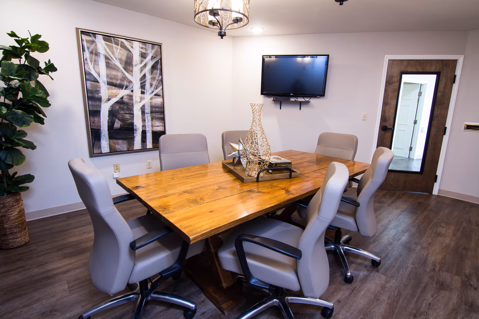 A modern conference room with a wooden table surrounded by six gray office chairs on wheels. A decorative centerpiece sits on the table. The room has wood flooring, a large framed artwork of white trees on a dark background on one wall, a flat-screen TV mounted on another wall, and a wooden door with a full-length mirror. A potted plant is in the corner, and a ceiling light fixture hangs above the table.