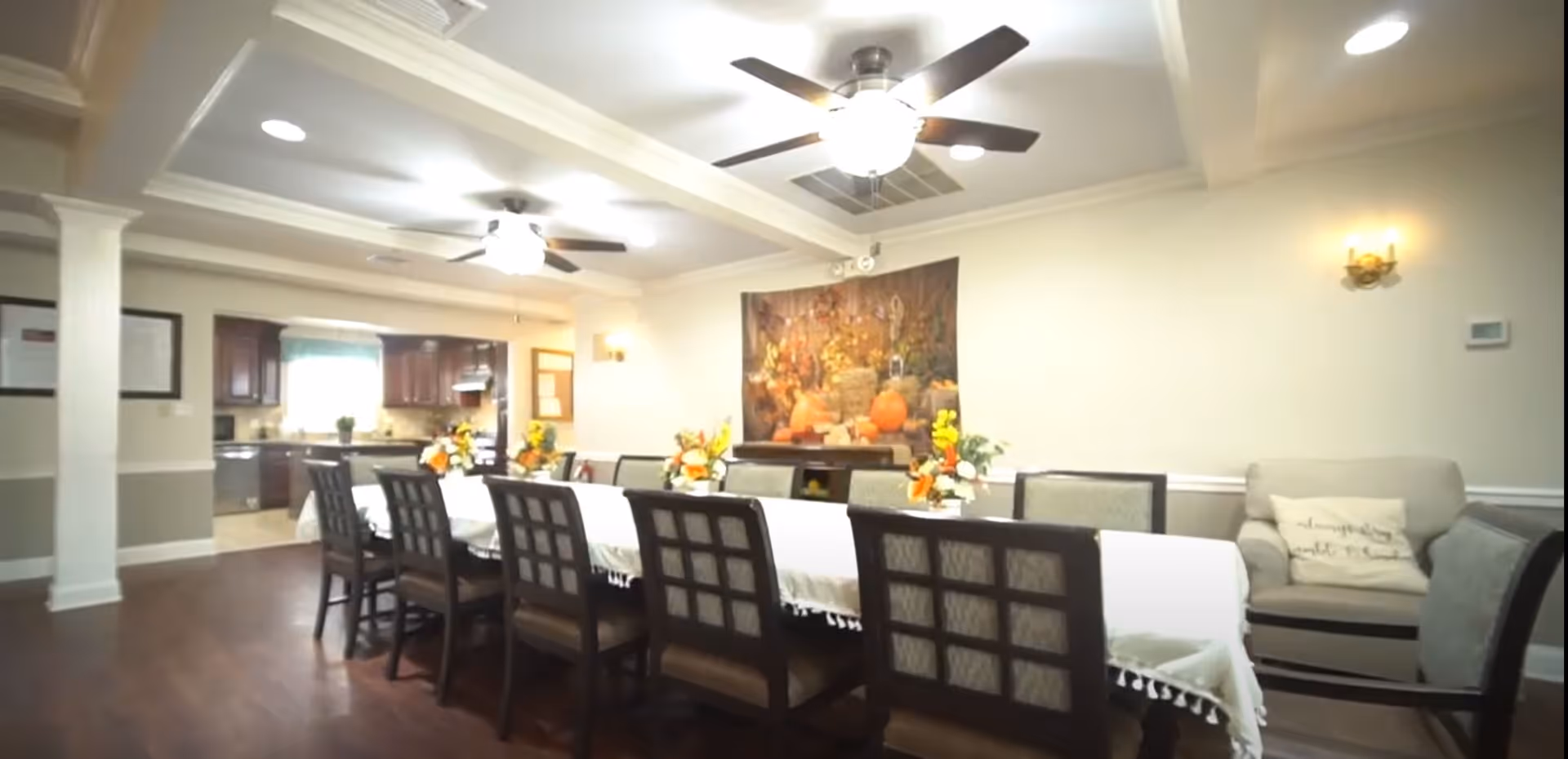 A spacious dining room with a long table covered by a white tablecloth and decorated with floral centerpieces. The room has wooden flooring, ceiling fans with lights, and a cozy seating area with a couch and chairs. The kitchen is visible in the background with dark wood cabinets and a window.