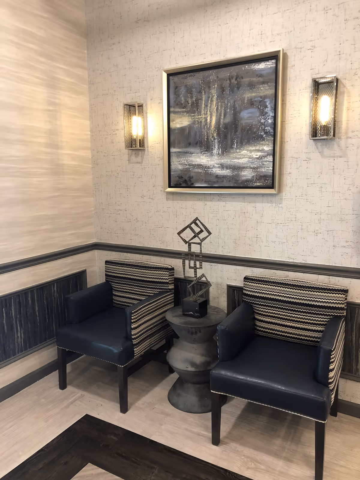 Two modern armchairs with patterned backs and dark blue seats are positioned around a small round pedestal table with a geometric metal sculpture on top. The wall behind features textured wallpaper, two wall sconces with warm lighting, and an abstract framed painting in shades of gray and white.