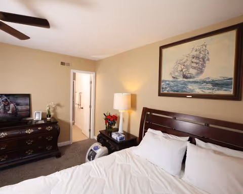 A bedroom in a senior living facility with a neatly made bed featuring white bedding and pillows. A dark wooden headboard is visible, along with a nightstand holding a lamp, a box of tissues, and a vase with red flowers. On the wall above the bed is a framed painting of a sailing ship on the ocean. Across from the bed is a dark wooden dresser with a flat-screen TV on top, displaying a scene with a person in a diving suit. The room has beige walls and carpeted flooring. An open door leads to a bathroom with grab bars visible.