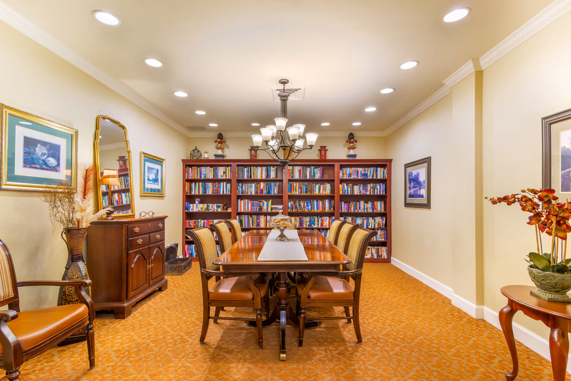 A well-lit common room with a long wooden table surrounded by chairs and bookshelves along the back wall.