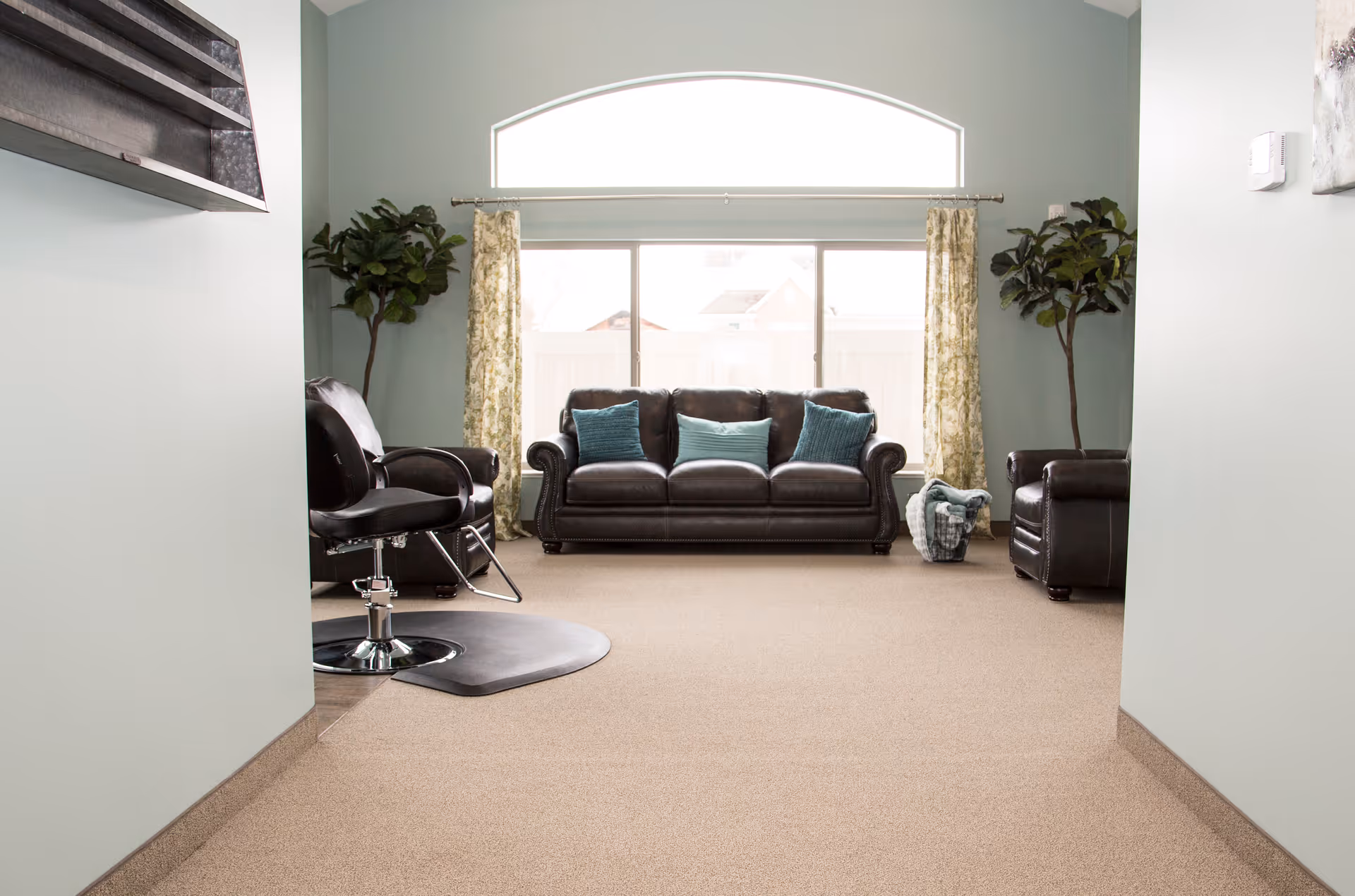A cozy living room with a large window featuring an arched top. The room has a dark leather sofa with three blue cushions, two matching leather armchairs, two tall potted plants, floral curtains, and a basket with blankets. There is also a black salon chair on the left side near the entrance.