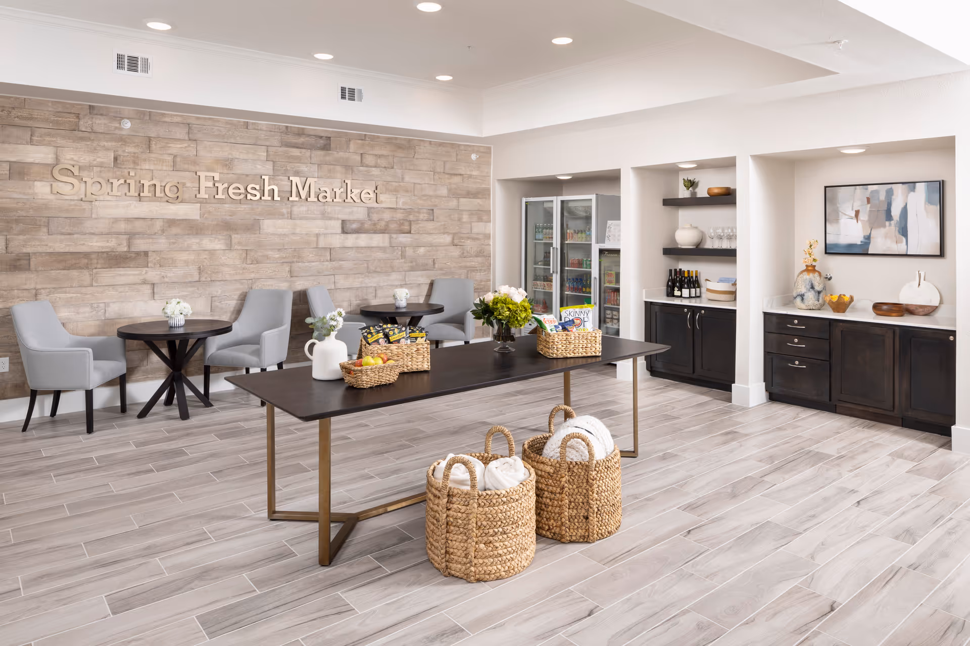 Interior view of a market area named 'Spring Fresh Market' with a wooden accent wall, two small round tables with gray chairs, a long table with baskets containing snacks and flowers, two woven baskets with rolled white towels, and built-in shelves and cabinets with decorative items and a refrigerator.