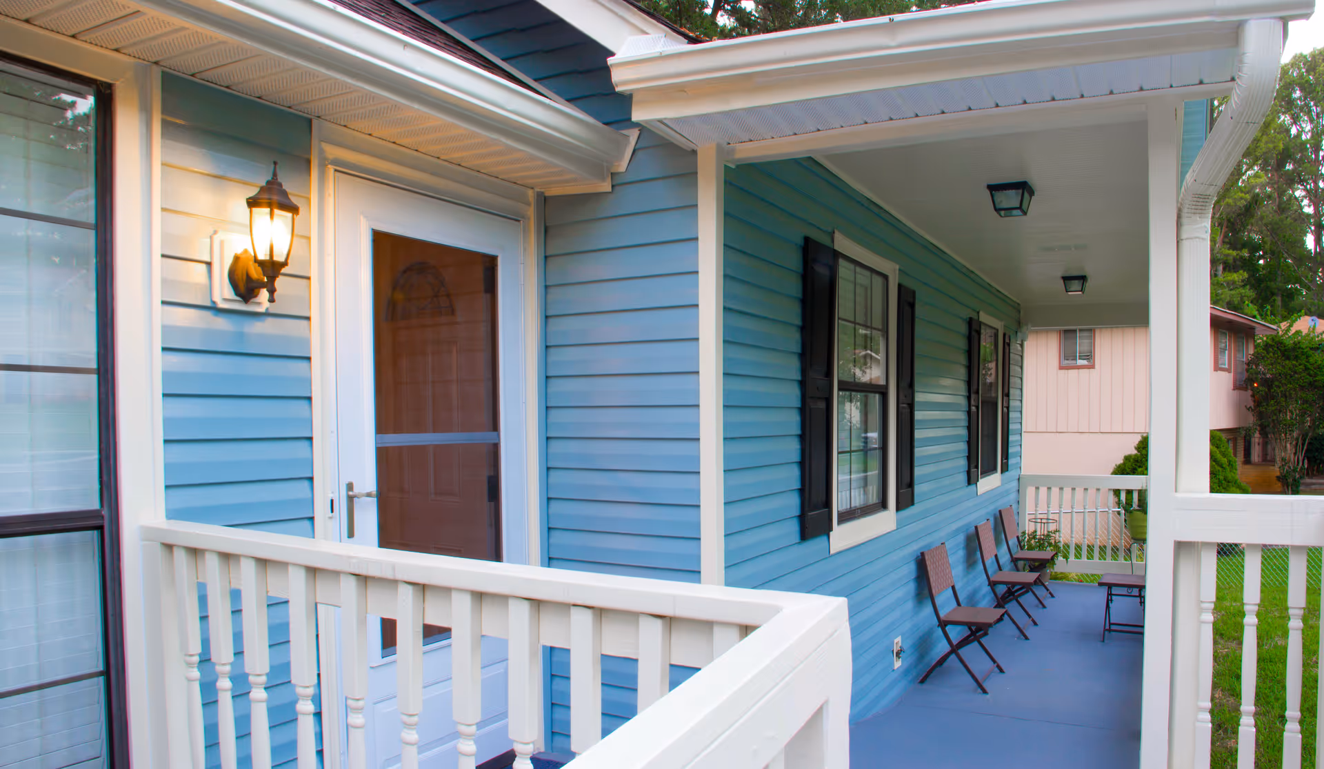 A covered front porch of a residential home with blue siding and white trim. The porch has a white railing, several brown folding chairs lined up against the wall, two black shuttered windows, and a lit wall-mounted lantern next to a white door with a glass storm door. The porch floor is painted blue and there are ceiling lights overhead.