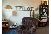 A cozy interior living space featuring a plush brown recliner chair, a tall floor lamp, a small side table with a candle, and a black shelving unit filled with decorative items and storage baskets. The walls are light-colored with framed pictures and a decorative fan hanging.