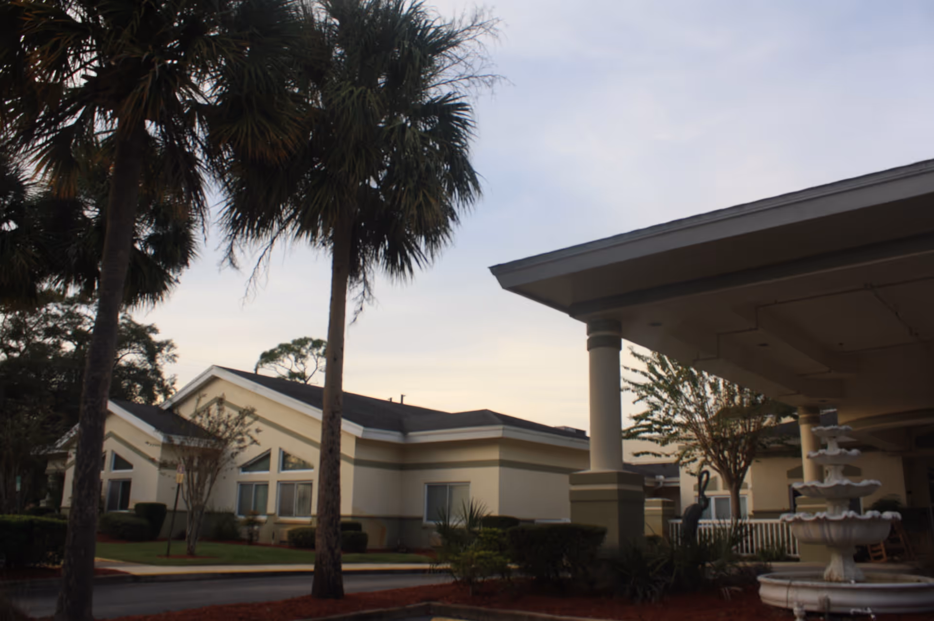 Exterior view of a senior living facility building with palm trees in the foreground and a multi-tiered white fountain near the entrance under a covered driveway at dusk.