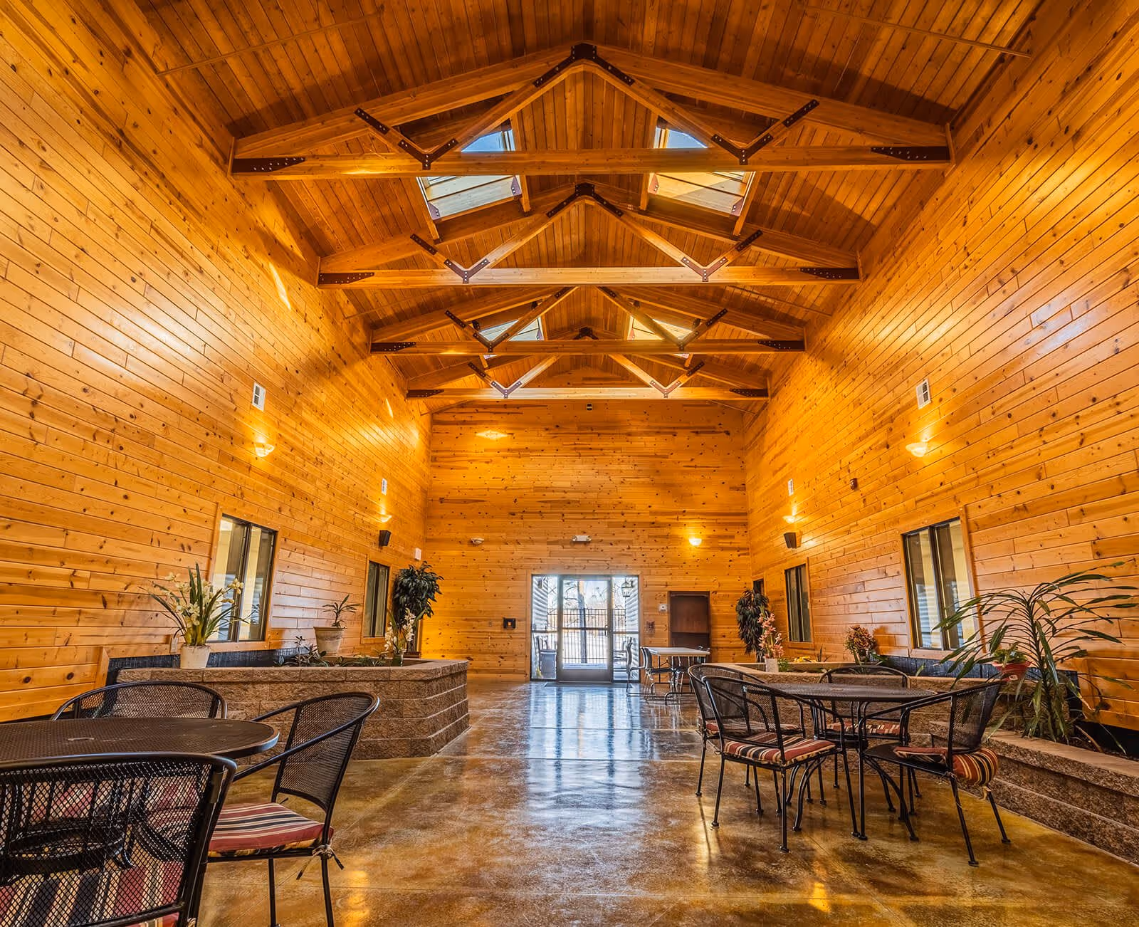 Interior of a spacious room with wooden walls and ceiling featuring exposed beams and skylights. The room has polished concrete floors and several metal tables and chairs with striped cushions. There are potted plants along the walls and a glass door at the far end letting in natural light.