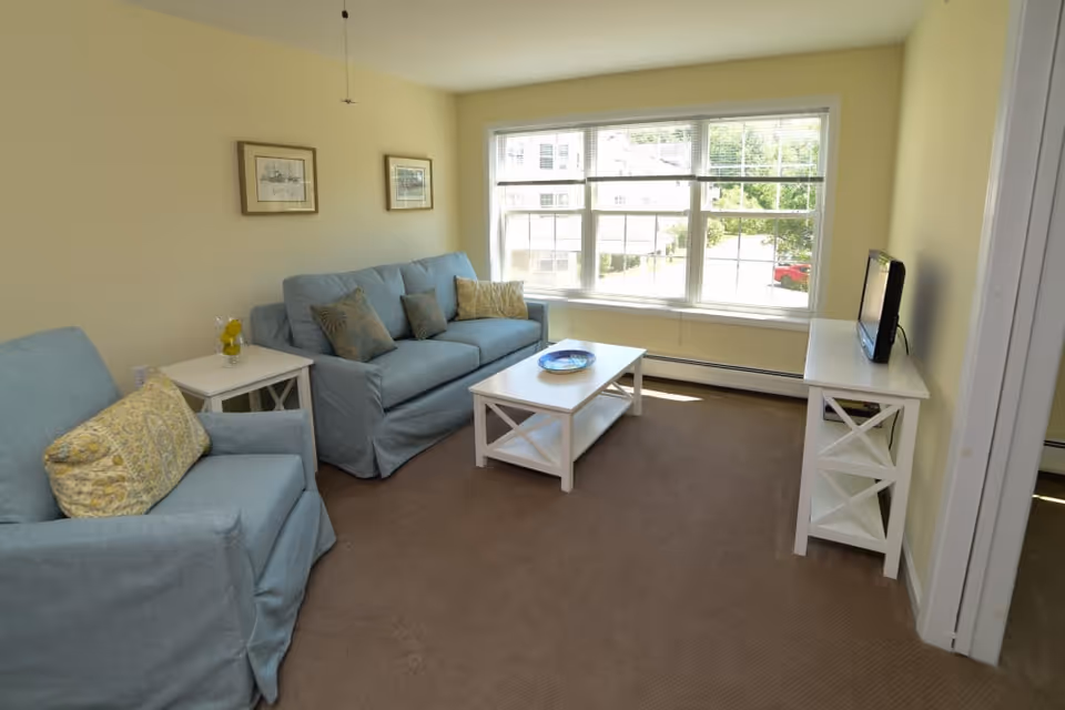 A cozy living room with light yellow walls and a large window letting in natural light. The room features a blue upholstered sofa and armchair with patterned cushions, a white coffee table with a decorative plate, a matching white side table with a glass jar containing lemons, and a white TV stand holding a small flat-screen television.