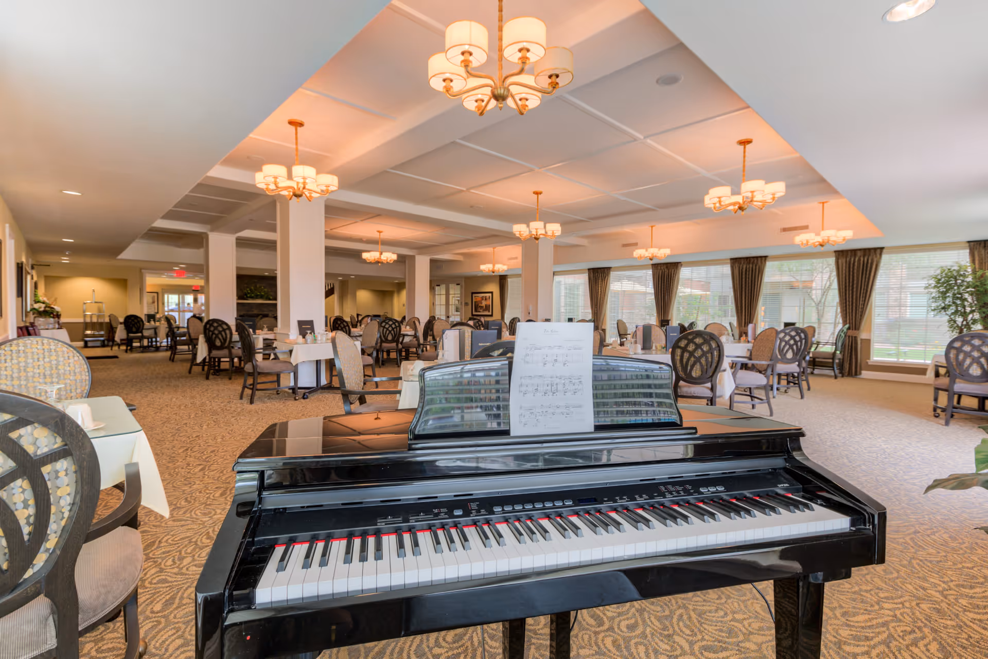 Interior view of a senior living facility dining room with multiple tables and chairs arranged neatly. A black grand piano with sheet music is prominently placed in the foreground. The room is well-lit with multiple chandeliers hanging from the ceiling and large windows with curtains allowing natural light to enter.