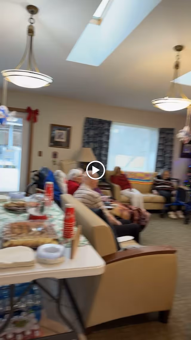 A common area in a senior living facility with several elderly people seated on couches and chairs. A table in the foreground holds snacks, red disposable cups, and bottled water. The room is well-lit with ceiling lights and skylights, and there are windows with curtains in the background.