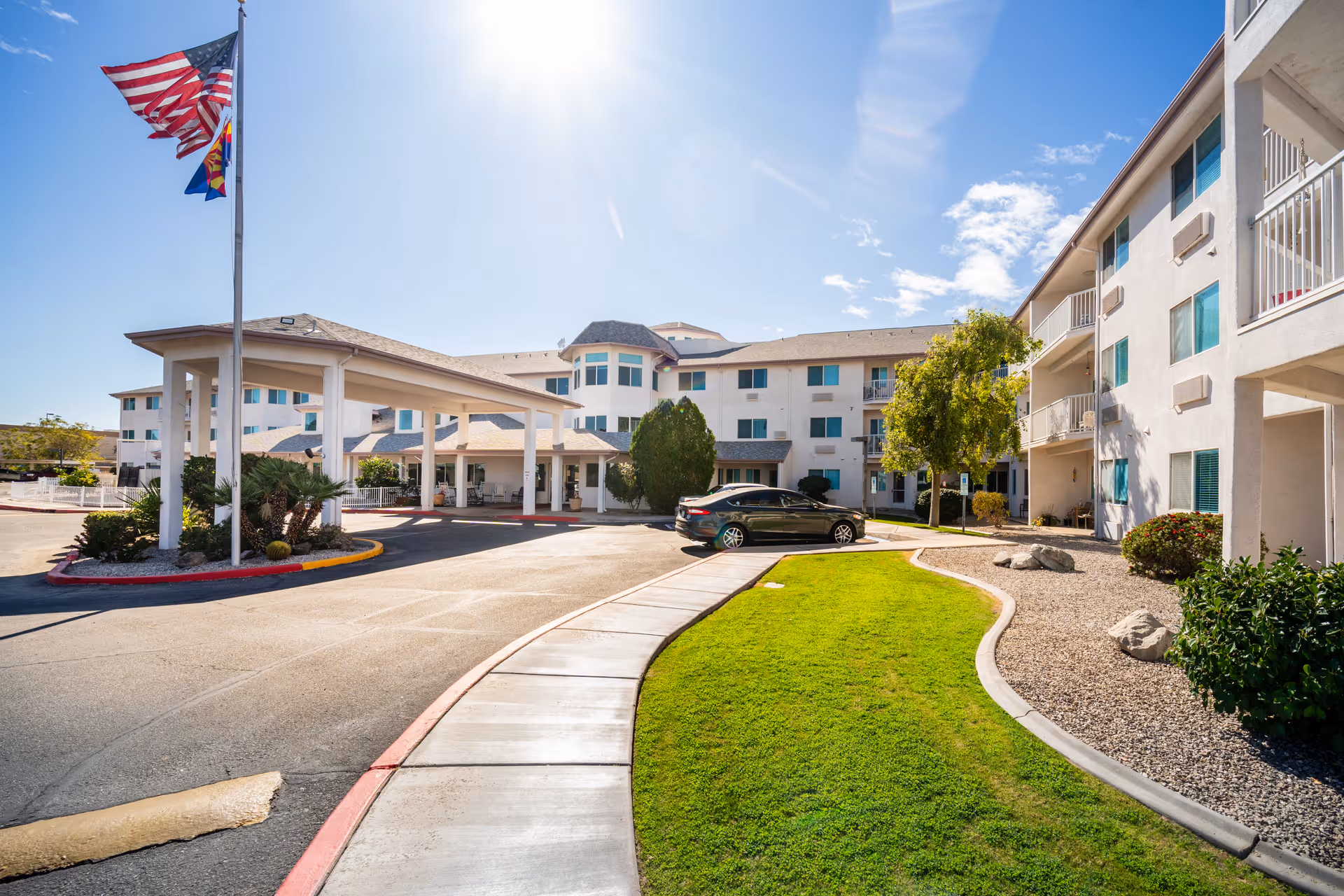 Exterior view of Desert Rose Retirement Community showing a multi-story building with balconies, a covered entrance, a driveway, a parked car, green lawn, and American and Arizona state flags on flagpoles under a clear blue sky.