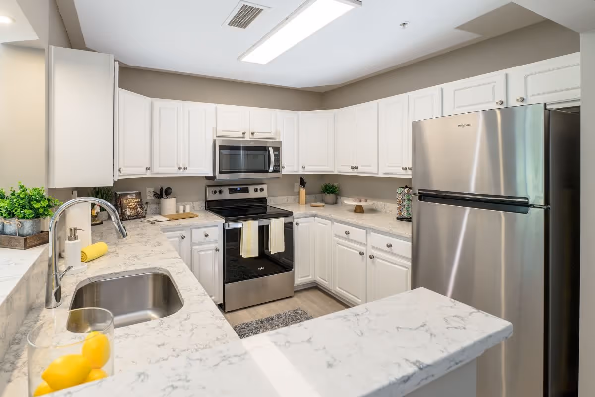 A modern kitchen with white cabinets, marble countertops, a stainless steel refrigerator, oven, and microwave. The kitchen features a sink with a chrome faucet, a small plant, a cutting board, and a container with lemons on the counter.