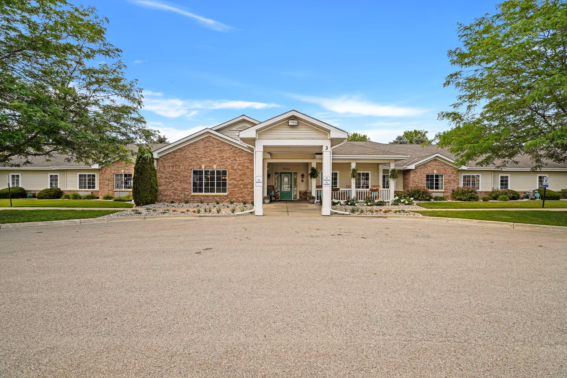 Front entrance of a single-story brick-and-siding senior living building with a covered portico, circular driveway, lawns, and trees.