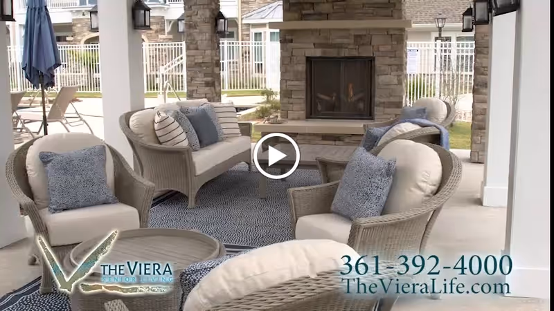 Outdoor covered patio area with wicker seating including chairs and a loveseat with cushions and pillows arranged around a coffee table. There is a stone fireplace in the background and a blue patterned rug on the floor. The area is part of a senior living facility with a fenced yard visible beyond the patio.