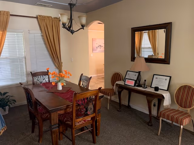 Cozy dining room with a wooden table and chairs, an orange floral centerpiece, a side console with a lamp and framed certificates, and a mirror on the wall.