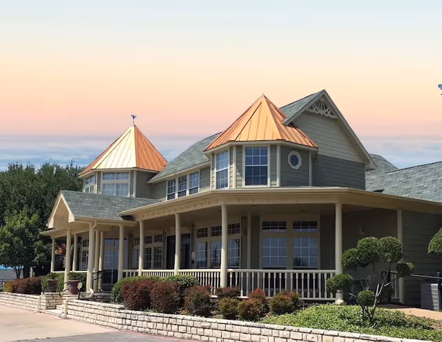 Exterior view of a senior living facility building with green siding, copper-colored turret roofs, large windows, a covered porch with white railings, and landscaped bushes and trees in front under a clear sky at sunset.