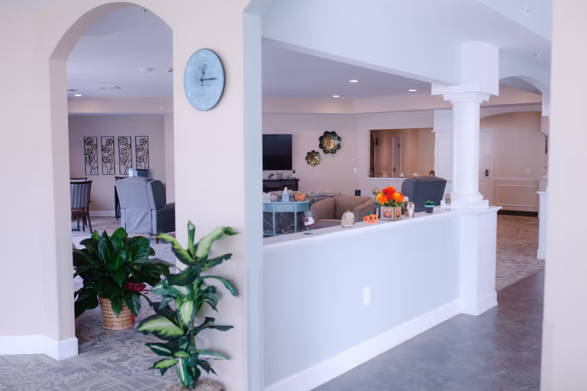 Open, airy senior living common area with seating, plants, columns and a TV visible beyond a half wall.