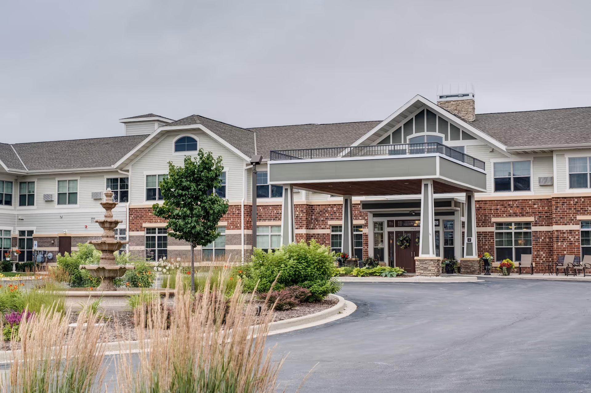 Front exterior of the Heritage West Allis senior living facility with a covered entryway, fountain, and landscaped driveway.