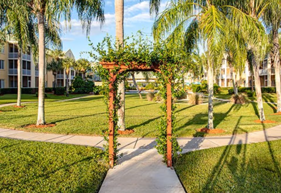 A sunny outdoor garden area with a wooden archway covered in green vines. The scene includes well-maintained grass, palm trees, and paved walkways. In the background, there are multi-story residential buildings with balconies.