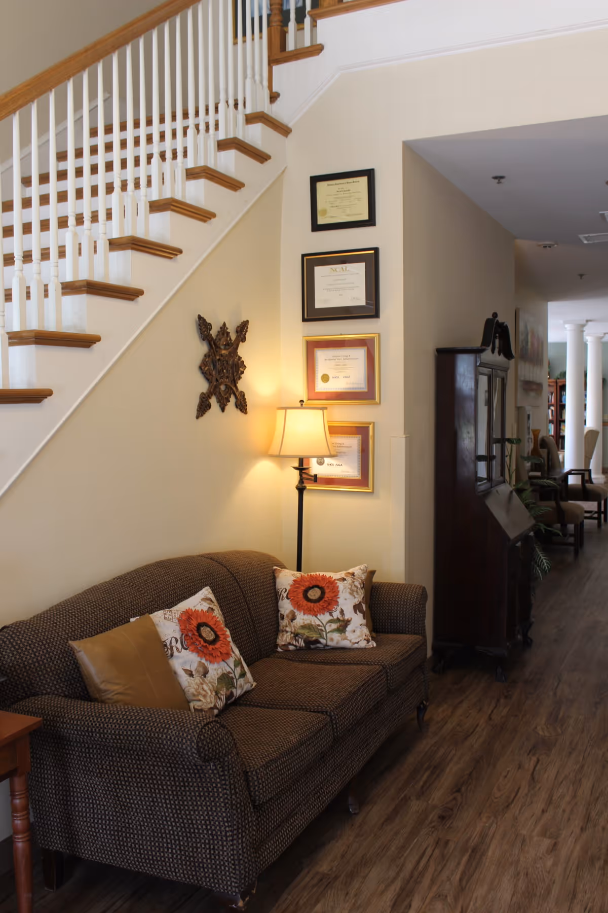 A cozy interior space featuring a brown patterned sofa with three decorative pillows, positioned under a staircase with wooden handrails and white balusters. A floor lamp beside the sofa illuminates framed certificates and a decorative wall piece on the wall. The room has wood flooring and leads into a hallway with additional seating and furniture visible in the background.