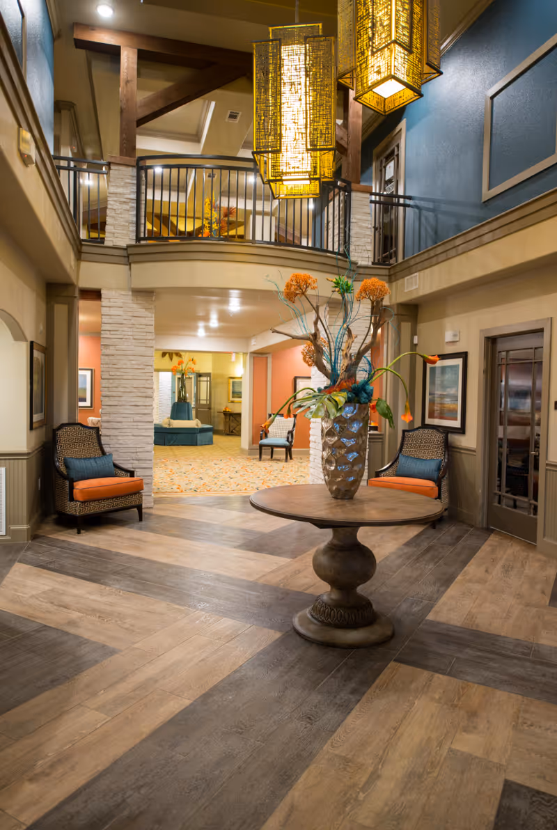 Interior view of a senior living facility lobby with a round wooden table in the center holding a decorative vase with orange and green floral arrangement. The space features wood and stone accents, two patterned armchairs with orange cushions on either side, a blue accent wall, and a second-floor balcony with black railing. Warm yellow hanging lanterns illuminate the area.